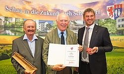 Drei Männer stehen auf einem Rasenfeld und halten eine Urkunde. Der Mann in der Mitte hält eine Trophäe. Sie lächeln. Ein Banner mit den Worten 'Schau in die Zukunft' steht hinter ihnen.