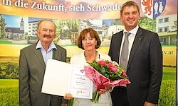 Drei Personen posieren für ein Foto mit einem Zertifikat und Blumen. Ein Mann und eine Frau in Anzügen stehen vor einem Banner.