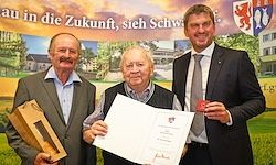 Drei Männer in Anzügen und Krawatten stehen vor einem Banner. Zwei halten Auszeichnungen und einer hält ein Zertifikat.