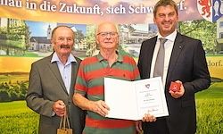 Drei Männer stehen nebeneinander. Der Mann in der Mitte hält ein Zertifikat. Der Mann auf der rechten Seite hält ein rotes Objekt. Der Mann auf der linken Seite hält eine kleine Tasche. Sie stehen vor einer grünen Kulisse.