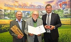 Drei Männer stehen vor einer mit Text bedruckten Kulisse, einer in der Mitte hält ein Zertifikat, während die anderen Holzplaketten halten.