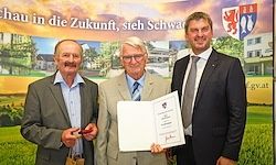 Drei Männer stehen auf einem Feld mit einem grünen Banner hinter ihnen. Der Mann in der Mitte hält ein Zertifikat und lächelt. Sie sind in Anzügen und Krawatten gekleidet.