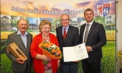 Vier Personen in formeller Kleidung stehen zusammen, zwei Männer halten ein Zertifikat und einen Blumenstrauß, eine Frau lächelt mit Blumen und ein Hintergrund mit einem Banner.