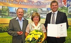 Drei Menschen stehen unter einem Banner. Zwei Männer in Anzügen und eine Frau, die einen Strauß gelber Blumen hält. Der Mann auf der rechten Seite hält ein Zertifikat.