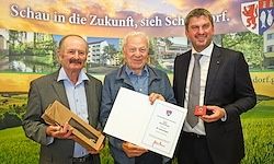 Drei Männer stehen vor einem Hintergrund mit einem Banner. Der Mann links hält eine Box, der Mann in der Mitte hält ein Zertifikat und der Mann rechts hält ein rotes Objekt. Sie alle lächeln.