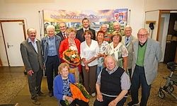 Eine Gruppe von Menschen, sowohl Männer als auch Frauen, steht für ein Foto vor einem Banner. Sie sind alle formell gekleidet, einige halten Blumen. Der Mann in der Mitte sitzt auf einem Stuhl.