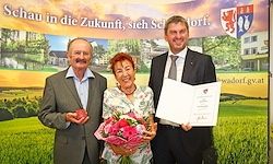 Drei Menschen stehen vor einem Banner und einem Grasfeld. Ein Mann hält ein Papier und Blumen. Ein anderer hält ein Papier und einen kleinen roten Gegenstand. Die Frau hält Blumen.