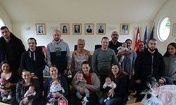 Eine Gruppe von Erwachsenen und Kindern posiert für ein Foto in einem Raum mit Bilderrahmen und Flaggen. Einige halten Babys.