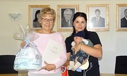 Zwei Frauen stehen vor einer Wand mit gerahmten Bildern. Eine hält ein eingewickeltes Baby und ein Buch, die andere ein eingewickeltes Geschenk.
