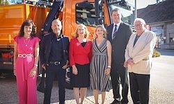 Sechs Menschen stehen vor einem orangefarbenen Lastwagen. Drei Frauen und drei Männer. Die Männer sind geschäftlich gekleidet und die Frauen lässig.
