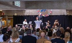 Eine Gruppe von Menschen schaut sich eine Aufführung auf der Bühne an. Die Bühne hat ein Podium, ein Klavier und eine Geige. Das Publikum sitzt auf Stühlen. Ein Mann steht auf dem Podium, und zwei Frauen spielen Geige.