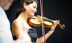 Eine Frau spielt Geige in einem dunklen Raum. Sie trägt ein ärmelloses Kleid und hält die Geige mit beiden Händen. Eine andere Person steht hinter ihr.