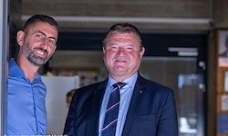Zwei Männer stehen nebeneinander und lächeln. Einer trägt ein blaues Hemd und der andere einen blauen Anzug mit Krawatte. Im Hintergrund befindet sich eine Glastür und ein Kühlschrank.