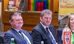 Zwei Männer in formellen Anzügen und Krawatten sitzen nebeneinander bei einer Versammlung, lächeln und schauen nach vorne. Eine Frau in einem weißen Hemd sitzt neben ihnen, und sie sind vor einem bunten Hintergrund mit einem Banner.
