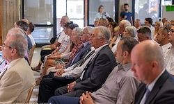 Eine Gruppe von Menschen in formeller Kleidung sitzt in Reihen in einem Auditorium, einige mit Brille. Sie schauen nach vorne und hören möglicherweise einem Redner zu.