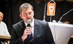 Ein Mann in einem dunklen Anzug hält ein Mikrofon und hält vermutlich eine Rede bei einer Veranstaltung mit einem Podium und Mikrofonen.