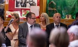 Eine Gruppe formell gekleideter Personen, darunter ein Mann mit Brille, sitzen in einem Sitzungssaal mit Flaggen und Dekorationen im Hintergrund.