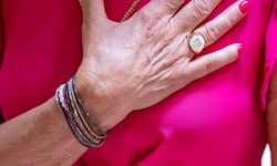 Eine ältere Frau mit roten Nägeln berührt ihre Brust mit der Hand. Sie trägt ein rosa Oberteil und mehrere Armbänder am linken Handgelenk.