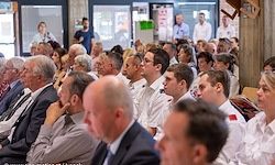 Eine Gruppe von Menschen in weißen Uniformen, wahrscheinlich medizinisches Personal, sitzt in einem Raum. Einige tragen Brillen, Krawatten und Ausweise. Ein Mann berührt sein Kinn. Im Hintergrund stehen und sitzen weitere Menschen.