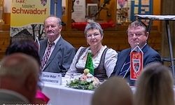Eine Gruppe von Menschen sitzt an einem Tisch mit einer Blumenarrangement und einem gelben Schild mit der Aufschrift 'Markt Schwadorf'. Zwei Männer und eine Frau lächeln und schauen in die Kamera. Der Mann auf der rechten Seite hält eine Fahne.