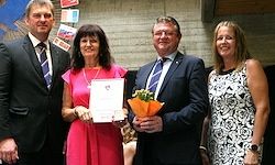 Vier Personen in formeller Kleidung posieren für ein Foto mit einer Auszeichnung und Blumen.