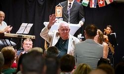 Ein älterer Mann mit Brille winkt einer Menge zu, während eine Person an einem Podium steht. Im Hintergrund sind Musikinstrumente zu sehen.