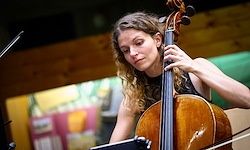 Eine Frau spielt Cello in einem Raum mit Holzwänden, verschwommenem Hintergrund und einer grünen Oberfläche hinter ihr.