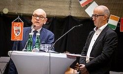 Zwei Männer stehen an einem Rednerpult mit Mikrofonen. Ein Mann hält eine Flagge mit einem Logo, der andere ein Buch. Hinter ihnen hängen Flaggen von der Decke.