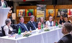 Mehrere Personen sitzen an einem langen Tisch, einige mit Getränken und Blumen. Sie scheinen an einer formellen Veranstaltung teilzunehmen. Ein Podium mit Mikrofon ist vorne aufgebaut.