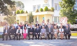 Eine Gruppe von Menschen in formeller Kleidung posiert für ein Foto vor einem Gebäude mit gelben Wänden und grünen Pflanzen obenauf.