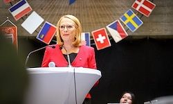 Eine Frau steht an einem Rednerpult und spricht in ein Mikrofon. Hinter ihr sind verschiedene Nationalflaggen aufgehängt. Sie trägt eine rote Jacke.