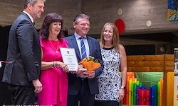 Eine Gruppe von Menschen steht bei einer Veranstaltung nebeneinander. Eine Frau in Pink und ein Mann im Anzug halten ein Zertifikat. Sie alle lächeln für die Kamera.