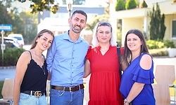 Vier Menschen stehen draußen vor einem Gebäude mit Bäumen und geparkten Autos. Drei Frauen und ein Mann. Der Mann trägt ein blaues Hemd und Jeans. Die Frauen tragen ein rotes Kleid, ein blaues Oberteil und ein schwarzes Top.