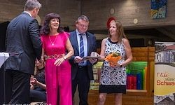 Ein Mann und eine Frau in formeller Kleidung stehen mit einem Blumenstrauß und überreichen einer Frau in einem rosa Kleid einen Preis. Dahinter steht ein weiterer Mann in Anzug und Krawatte mit einem Zertifikat. Die Umgebung umfasst eine Holzstruktur und ein Banner mit der Aufschrift 'F Life MARK SCHWA'.