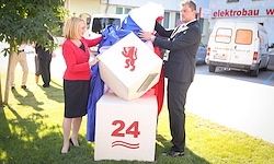 Ein Mann und eine Frau stehen vor einem würfelförmigen Objekt mit der Nummer 24 darauf, sie halten beide den blauen und weißen Stoff, der das Objekt bedeckt. Sie befinden sich in einem Außenbereich mit einem Gebäude und geparkten Autos im Hintergrund.