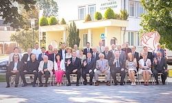 Eine große Gruppe formell gekleideter Personen posiert für ein Foto vor einem Gebäude mit gelber Fassade und Grünflächen. Im Hintergrund sind Bäume und ein klarer Himmel zu sehen.