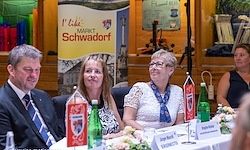 Drei Personen sitzen an einem Tisch mit Blumen und Flaschen. Ein Mann und zwei Frauen, alle lächeln. Im Hintergrund befindet sich ein Banner mit der Aufschrift 'Ich mag MARKT SCHWADORF'.