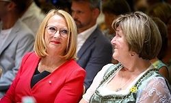 Zwei Frauen sitzen zusammen in einem überfüllten Raum. Die Frau auf der linken Seite trägt einen roten Blazer und eine Brille. Die Frau auf der rechten Seite trägt ein traditionelles bayerisches Kleid und hat eine gelbe Blume auf der Brust.