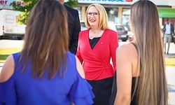 Drei Frauen stehen auf der Straße vor einem Geschäft. Die Frau in Rot lächelt und trägt eine Brille. Es sind Autos am Straßenrand geparkt.