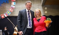Ein Mann in Anzug und eine Frau in rotem Blazer stehen nebeneinander und lächeln. Sie halten einen Blumenstrauß. Dahinter ist ein Musikinstrument zu sehen.