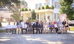 Eine Gruppe von Menschen in Anzügen und Kleidern posiert für ein Foto vor einem Gebäude mit Pflanzen und einer Flagge.