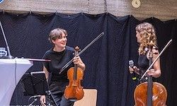 Zwei Frauen auf der Bühne, eine hält eine Geige und die andere ein Cello, lächeln und sehen sich an. Sie stehen auf einer Bühne mit schwarzem Hintergrund und einem Mikrofon.