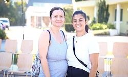 Zwei Frauen stehen vor einer Reihe von Stühlen und lächeln für ein Foto. Sie haben Taschen auf ihren Schultern. Dahinter stehen einige Bäume und ein Gebäude.