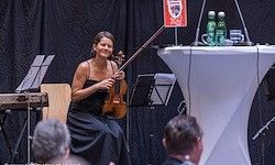 Eine Violinistin spielt auf der Bühne vor einem Publikum. Ein rotes Schild und zwei Flaschen stehen auf einem Podium hinter ihr.