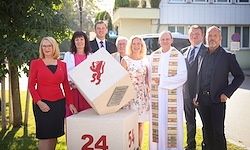 Eine Gruppe von Menschen steht draußen, einer hält eine weiße Box mit der Nummer 24. Bäume und ein Gebäude im Hintergrund.