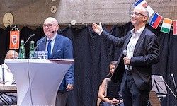 Zwei Männer in Anzügen stehen auf der Bühne; einer spricht in ein Mikrofon, der andere gestikuliert. Ein Rednerpult, Flaggen und ein sitzendes Publikum sind im Hintergrund.