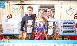 Drei Personen stehen mit Schürzendrueck mit Illustrationen eines Hundes, eines Maedchens und eines Mannes hinter einem Tisch. Ein Mann traegt ein Hemd mit einem Hund. Eine Frau traegt ein Kleid und ein Mann ein Hemd. Dahinter befinden sich Kartons und herzfoermige Dekorationen.