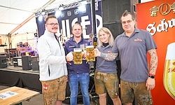 Vier Personen stehen vor einer Bühne und halten Biergläser in der Hand. Sie scheinen bei einer Outdoor-Veranstaltung zu feiern, mit einem Banner mit einem Bierglas im Hintergrund.