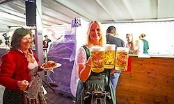 Zwei Frauen in traditioneller Kleidung halten Biergläser bei einem Festival. Eine Frau hält einen Teller mit Essen, während die andere zwei Gläser Bier hält.