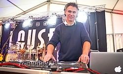 Ein Mann steht vor einer DJ-Anlage mit Laptop und Equipment auf der Bühne und lächelt. Hinter ihm ist ein Banner mit Text und Musikinstrumenten.